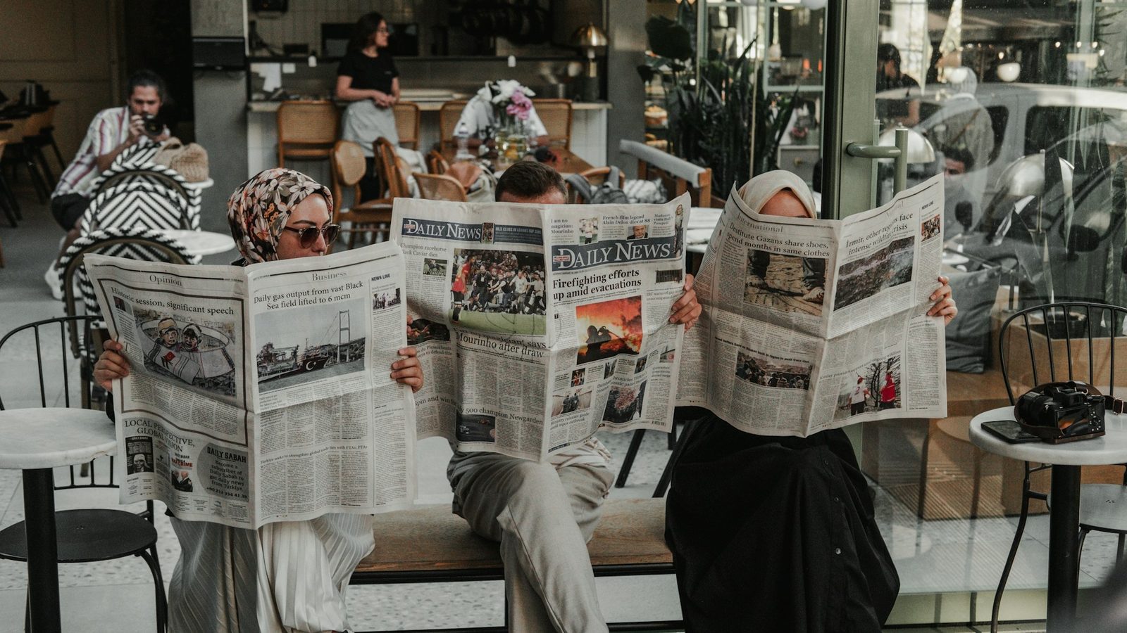 Tre personer leser aviser på en kafé – kildekritikk og nyheter